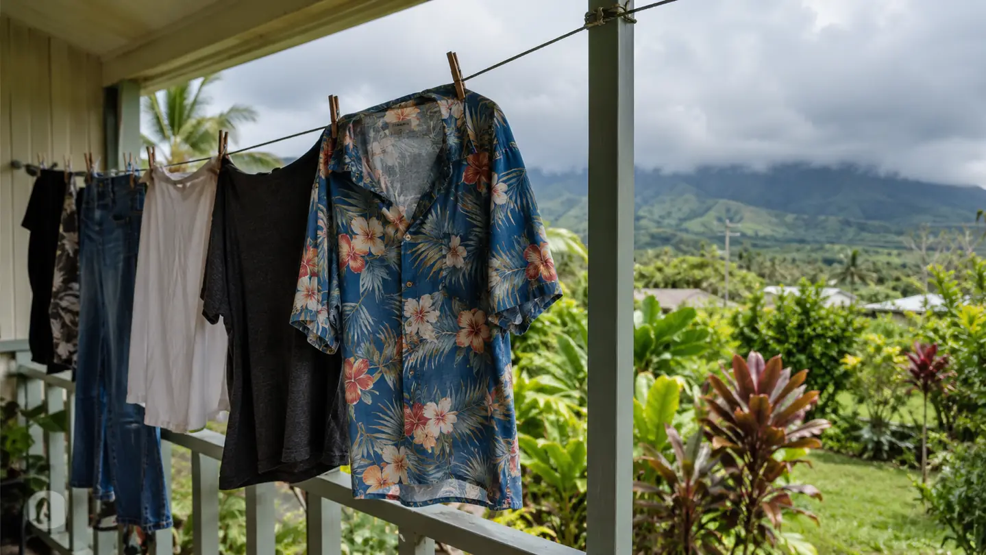 A short-sleeve Hawaiian shirt hanging on a clothesline on a lanai with tropical plants behind it